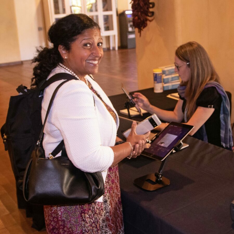 Image: One individual smiles at the camera while using a tablet, another person sits behind a table using another tablet