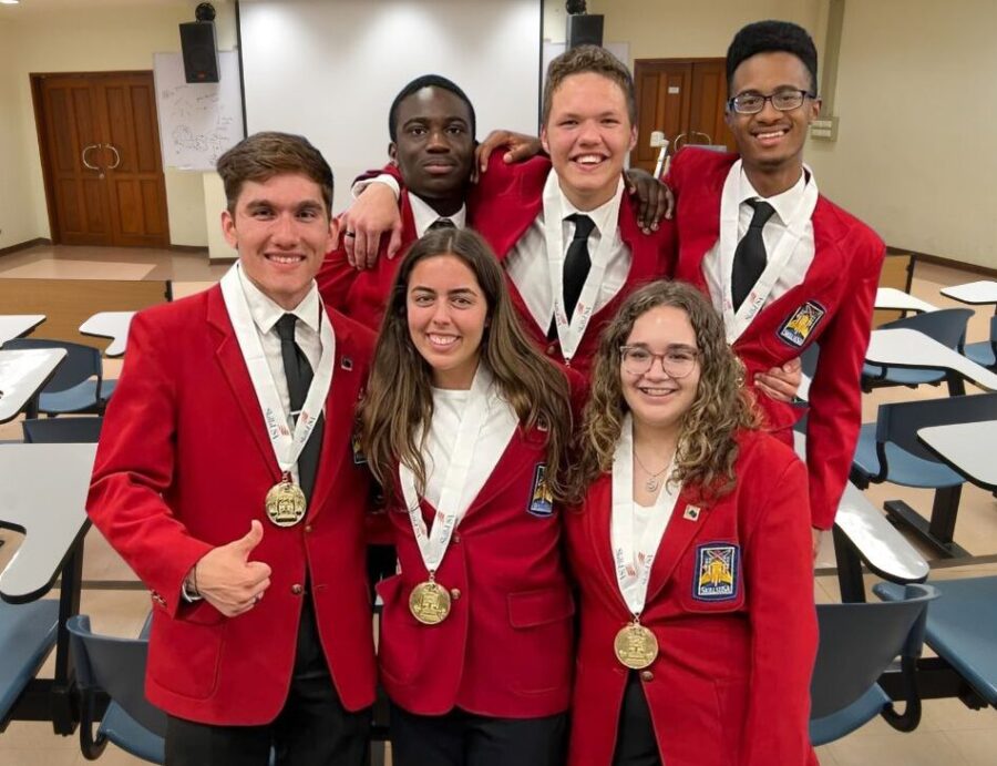 A groups of youth parliamentarians stand in a classroom wearing red blazers with awards medals.